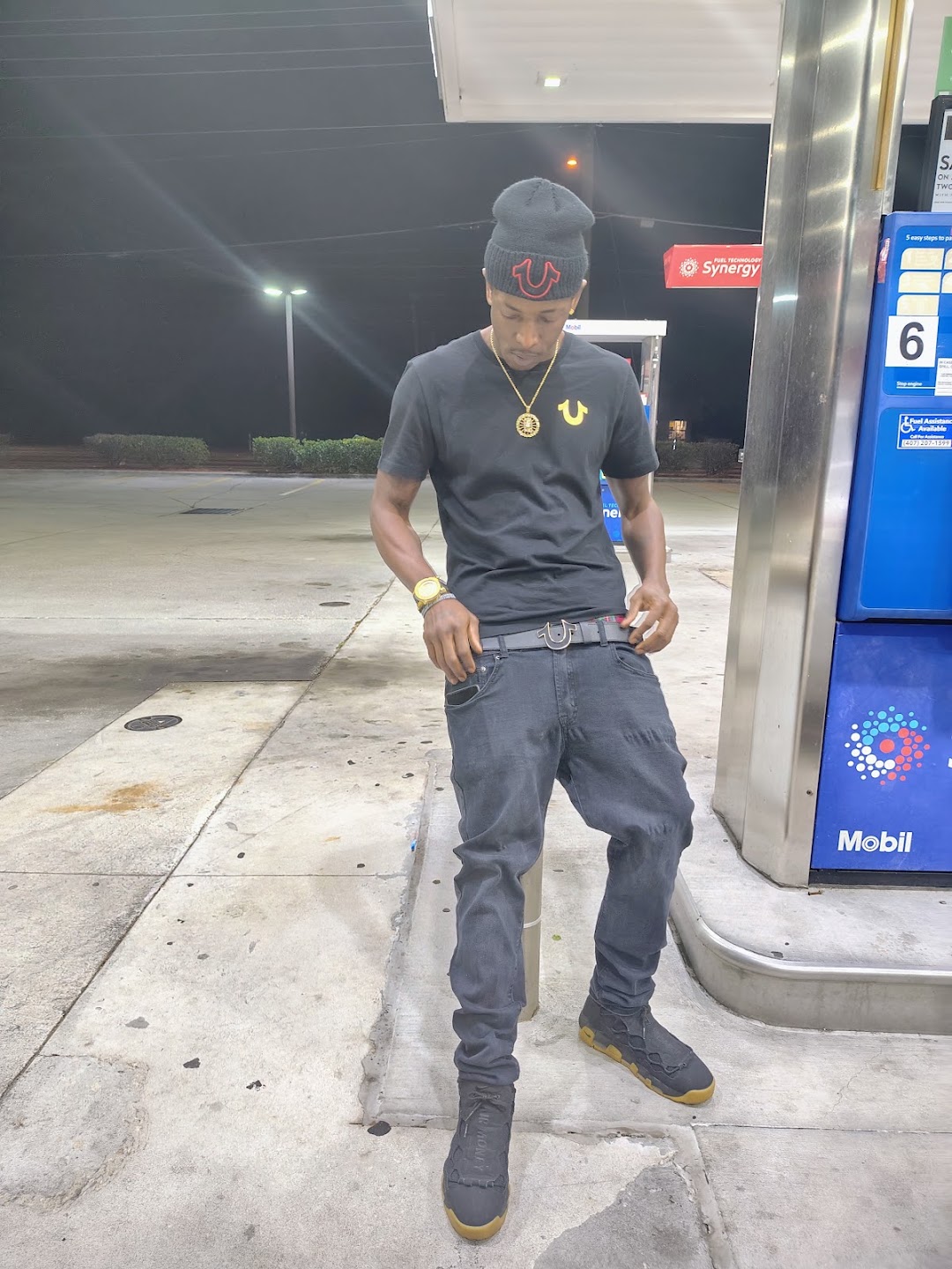 Twan G Young Gutta wearing a black True Religion shirt, beanie and belt at a gas station at night