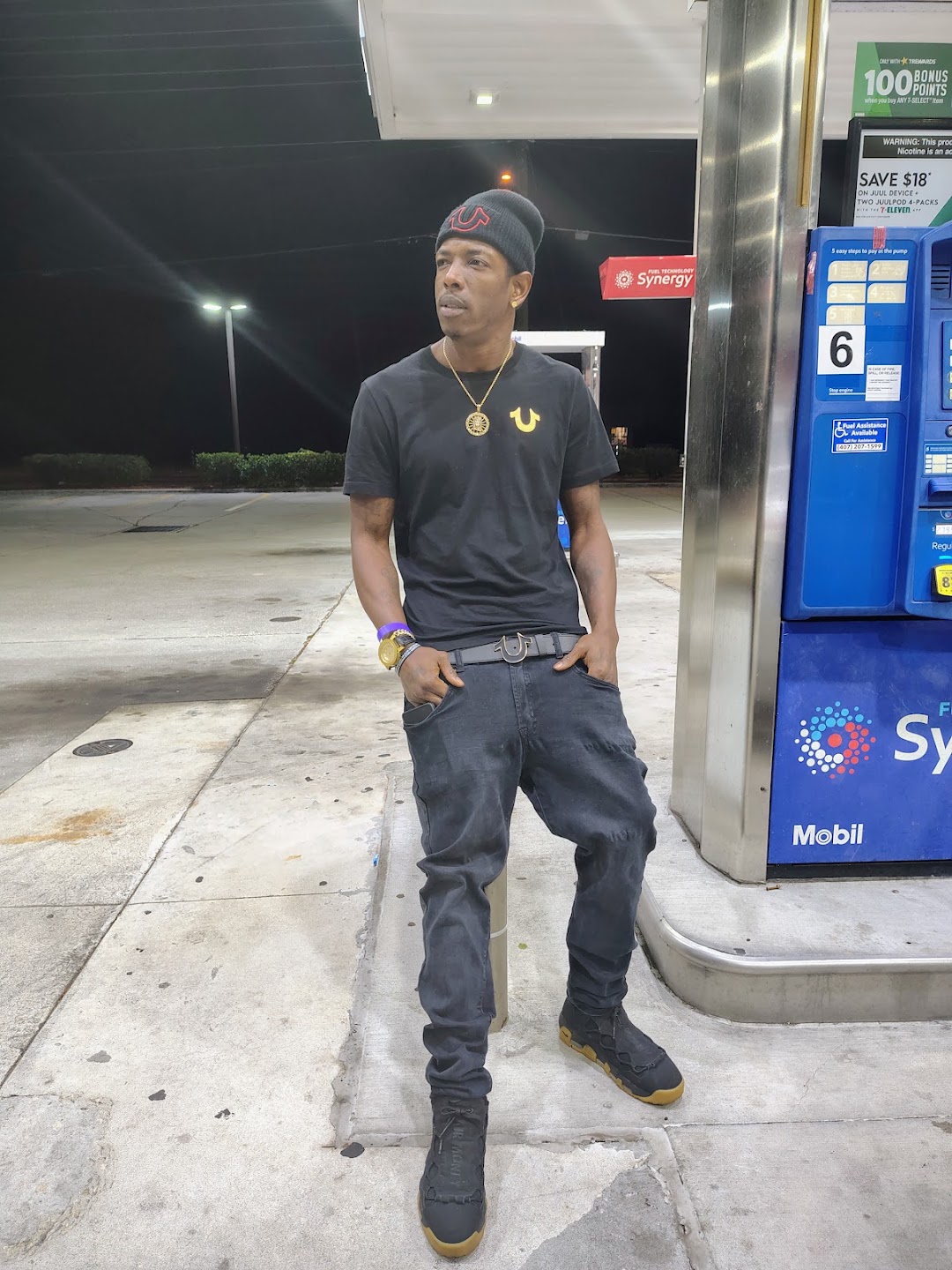 Full-length nighttime portrait of Twan G Young Gutta wearing a black True Religion shirt, belt and beanie