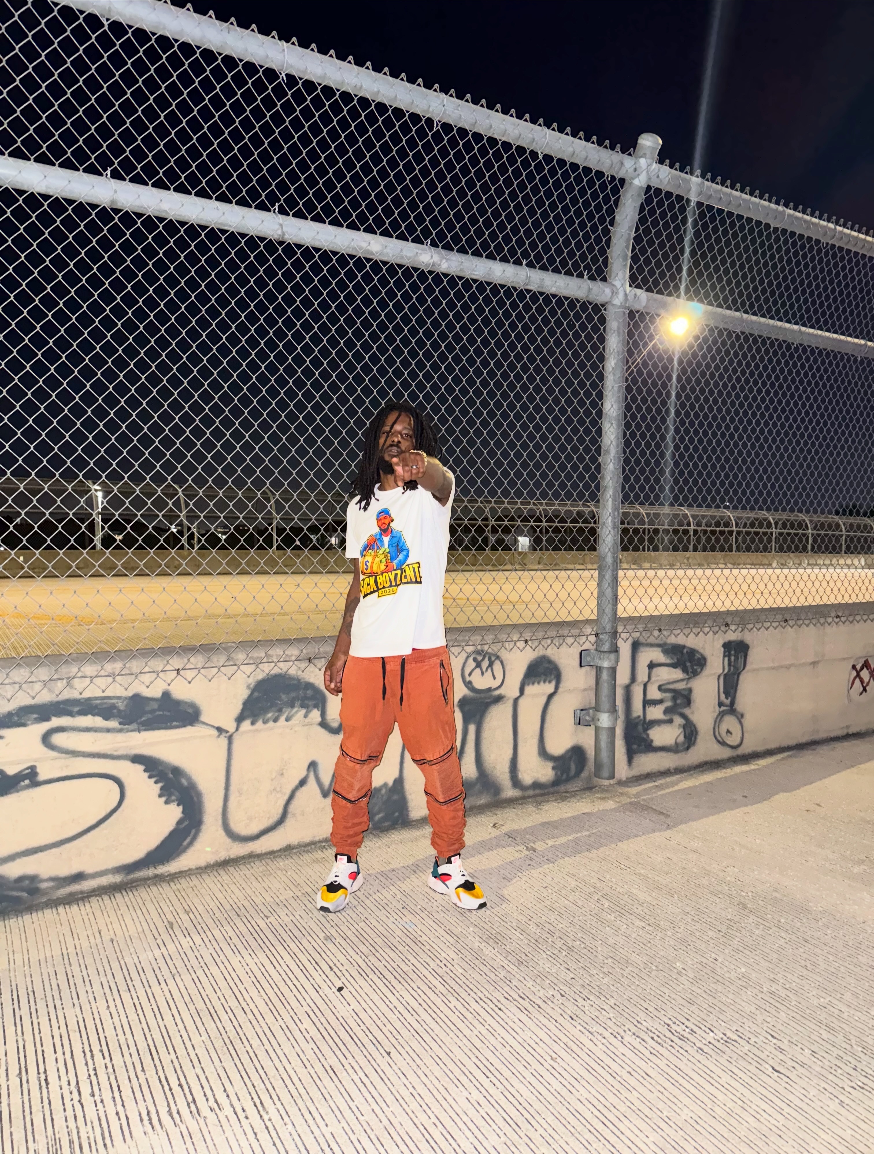 Chevy GC wearing a white Sack Boyz Entertainment 2026 T-shirt outdoors by a fence at night